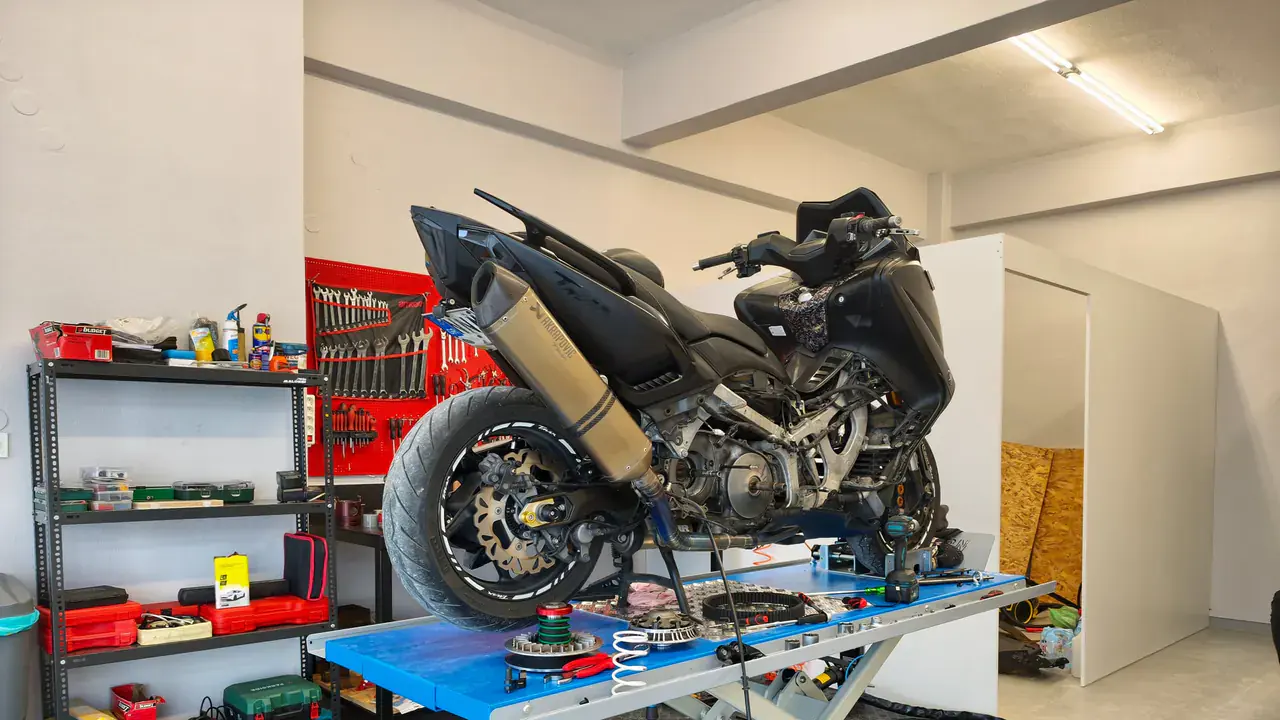 Μαύρο scooter σε μπλε ψαλιδωτό ανυψωτικό, με ανοιχτό κινητήρα και ανταλλακτικά πάνω στην πλατφόρμα.