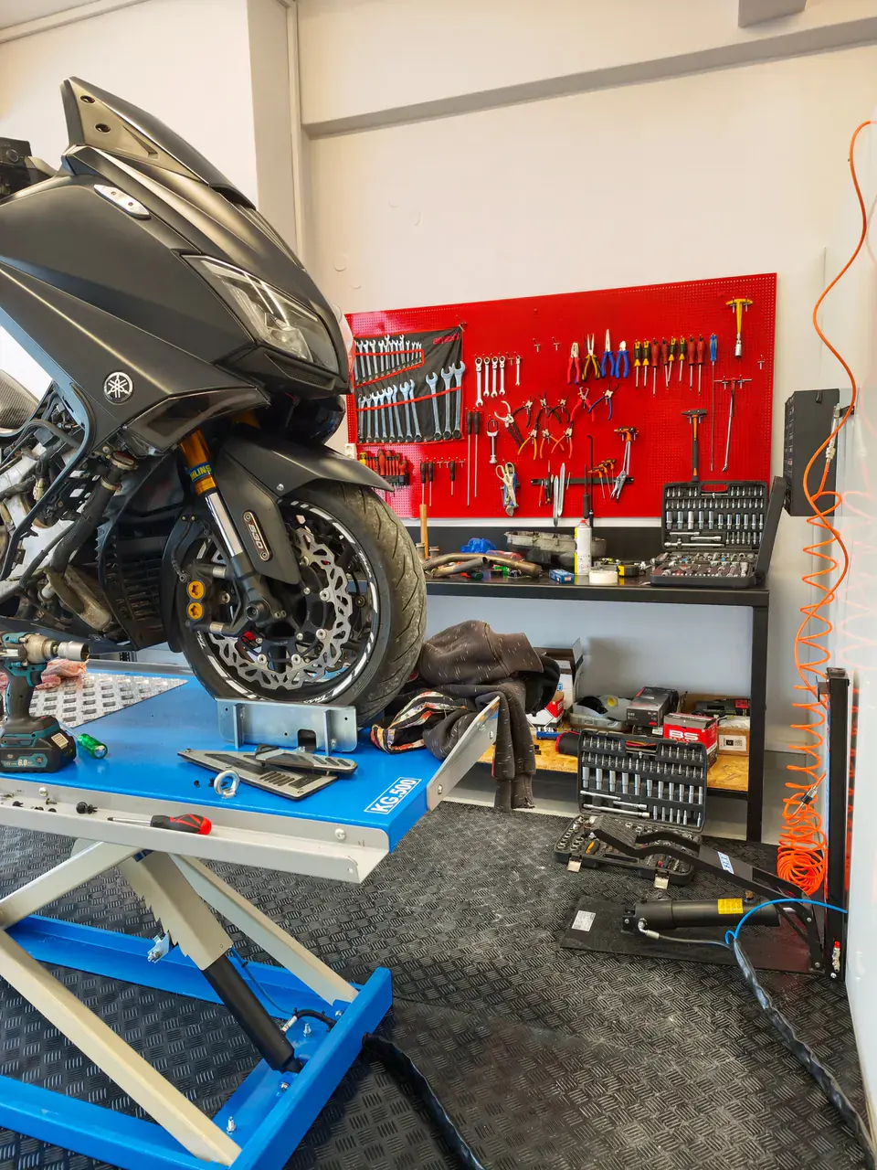 Μπροστινός τροχός scooter πάνω στο μπλε ανυψωτικό, με τον κόκκινο πίνακα εργαλείων στο βάθος.