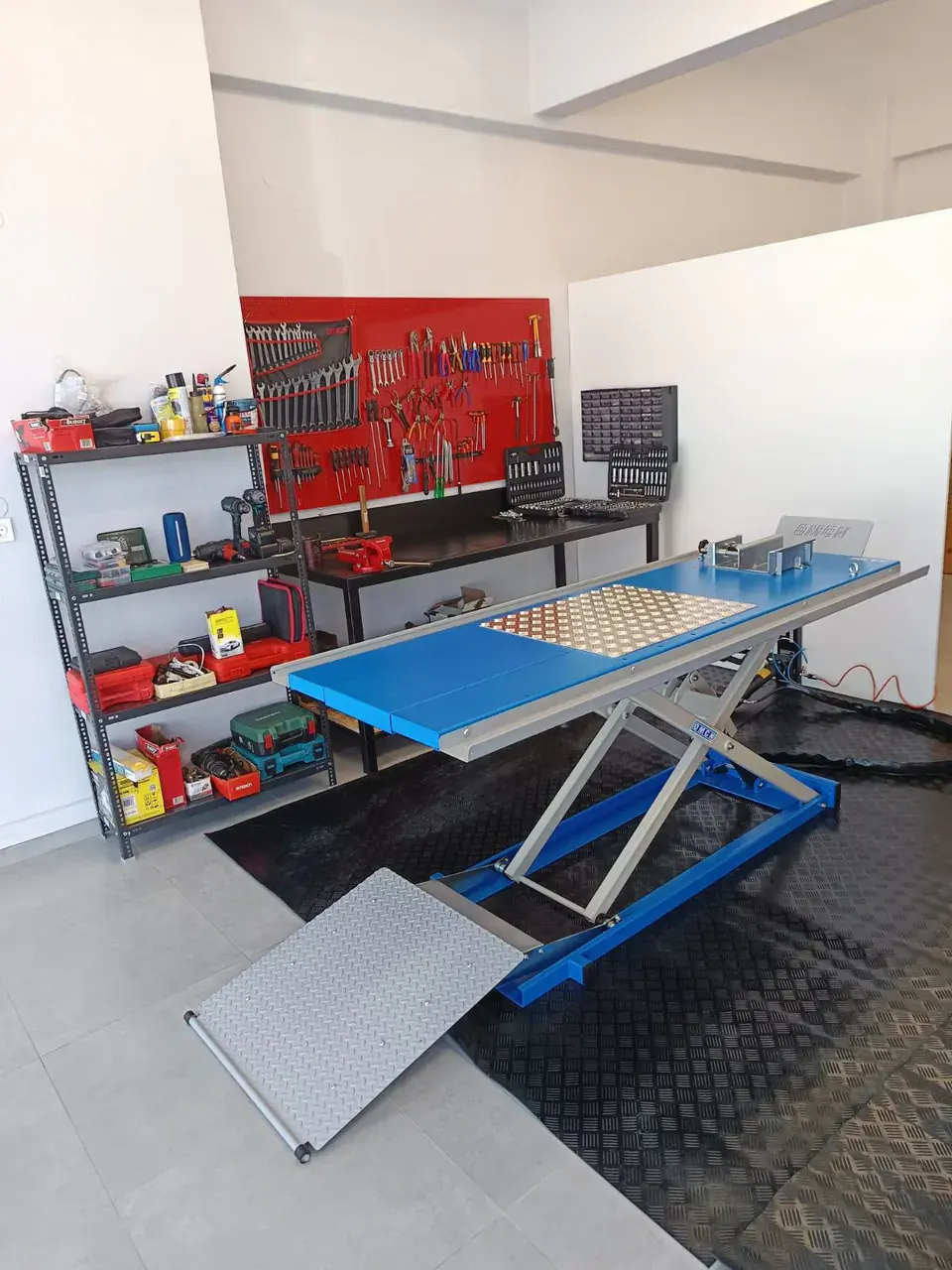Το μπλε ψαλιδωτό ανυψωτικό OMCN μπροστά από τον πάγκο και τον κόκκινο πίνακα εργαλείων.