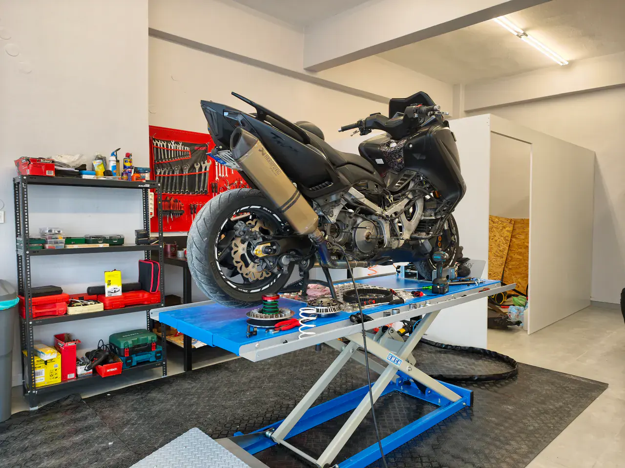 Μαύρο scooter στο μπλε ψαλιδωτό ανυψωτικό του συνεργείου, με τον κόκκινο πίνακα εργαλείων στο βάθος.