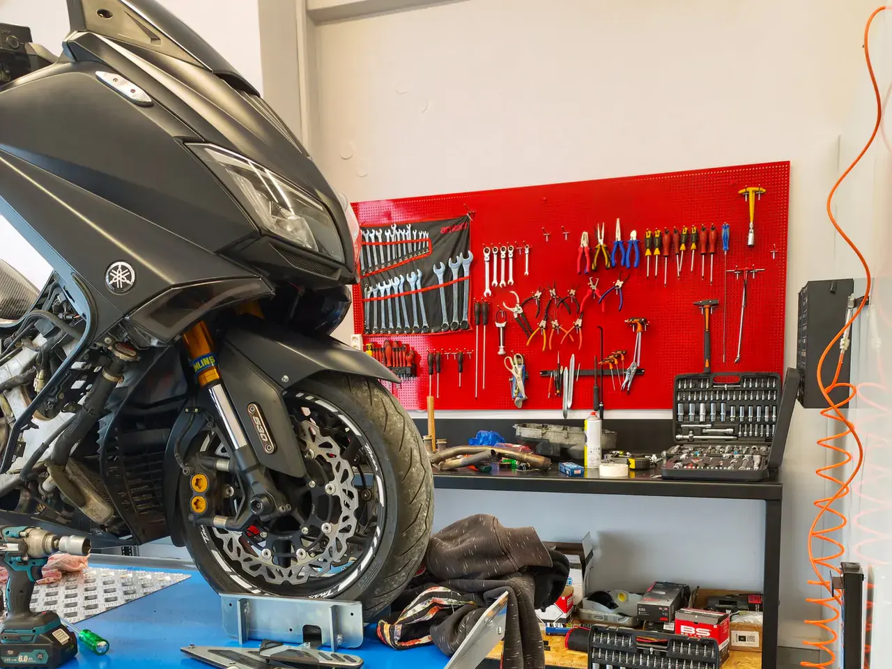 Μπροστινός τροχός scooter στο μπλε ανυψωτικό, με τον κόκκινο πίνακα εργαλείων στο βάθος.